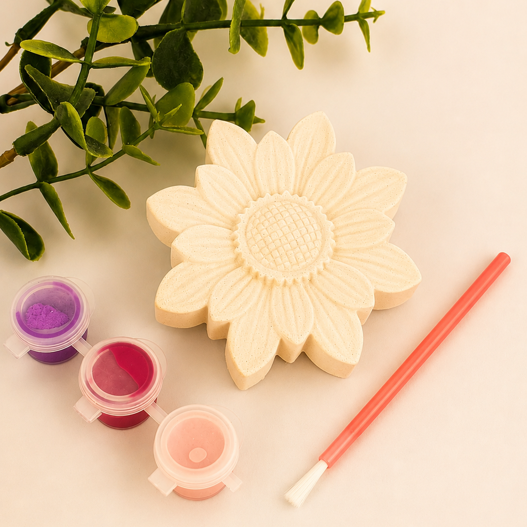 A flower-shaped craft painting kit on a light beige background, featuring a cream-colored flower mold, three small paint pots in purple, pink, and peach, a coral paintbrush, and green decorative leaves.